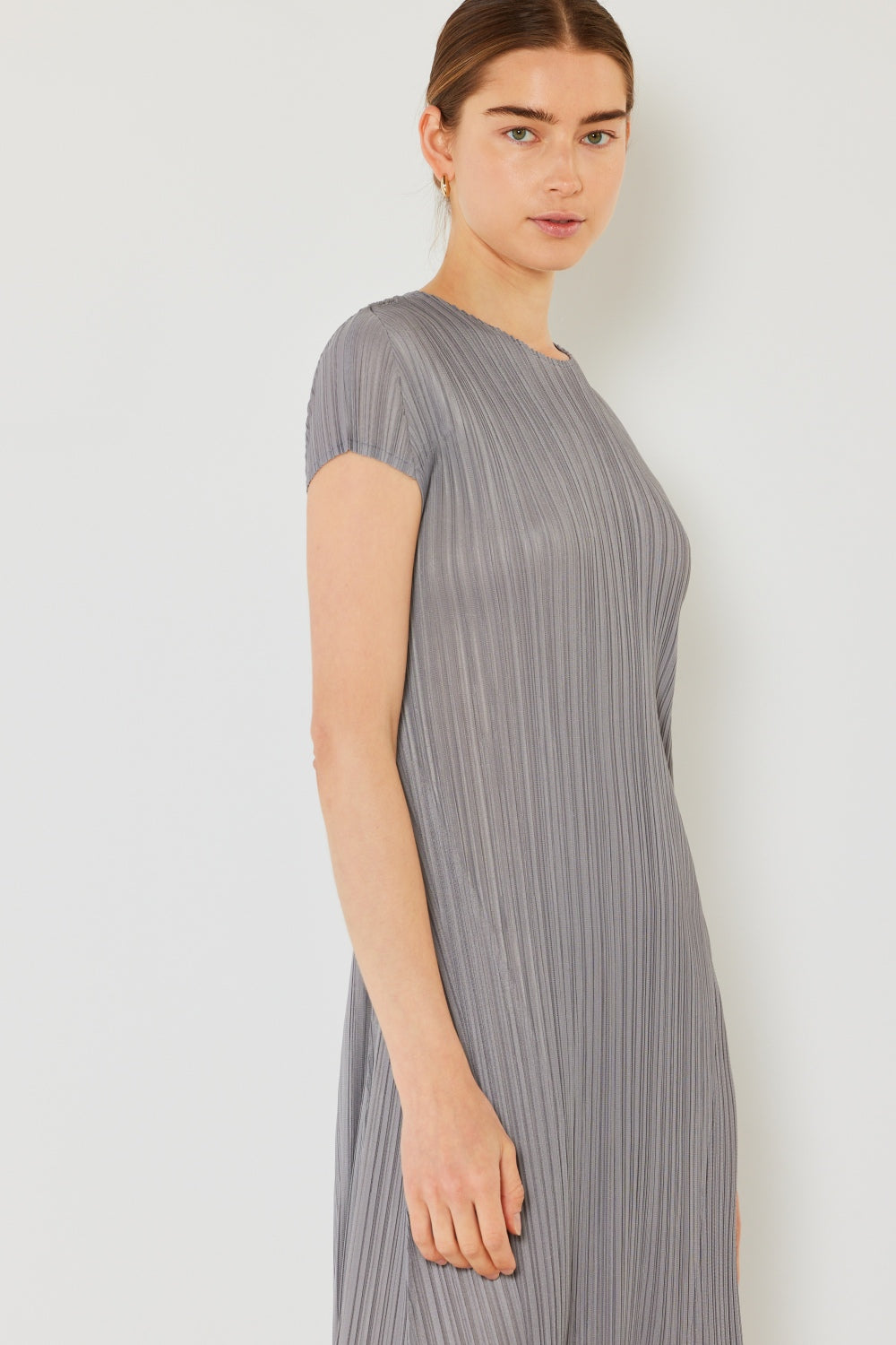 A woman with dark hair tied back wears the Marina West Swim Pleated Cap Sleeve A-Line Dress in light gray, exuding feminine charm. She stands against a neutral background, gazing softly at the camera.