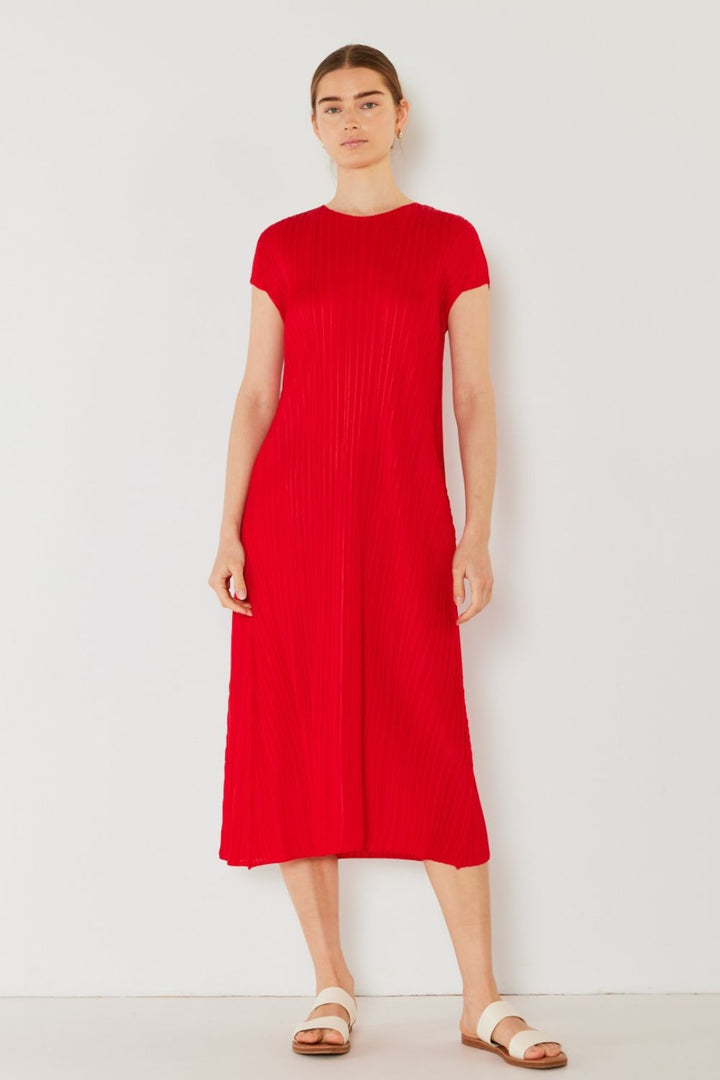 Against a simple backdrop, a person exudes feminine charm wearing the Marina West Swim Pleated Cap Sleeve A-Line Dress in red, paired with white sandals. Their long brown hair is tied back, and they wear a neutral expression.