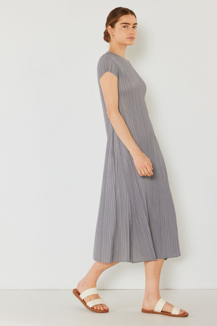 A person with brown hair tied back is wearing a Marina West Swim Pleated Cap Sleeve A-Line Dress in gray, paired with white sandals, exuding feminine charm. They stand sideways against a plain white background.