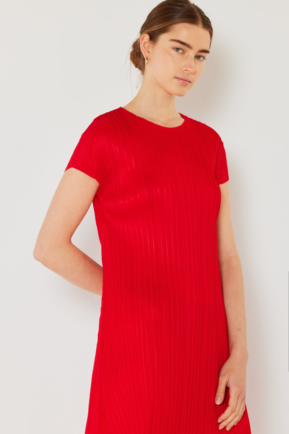 A person with brown hair tied back is wearing a red Marina West Swim Pleated Cap Sleeve A-Line Dress, exuding feminine charm. They stand against a light-colored wall, looking slightly to the side with a neutral expression.