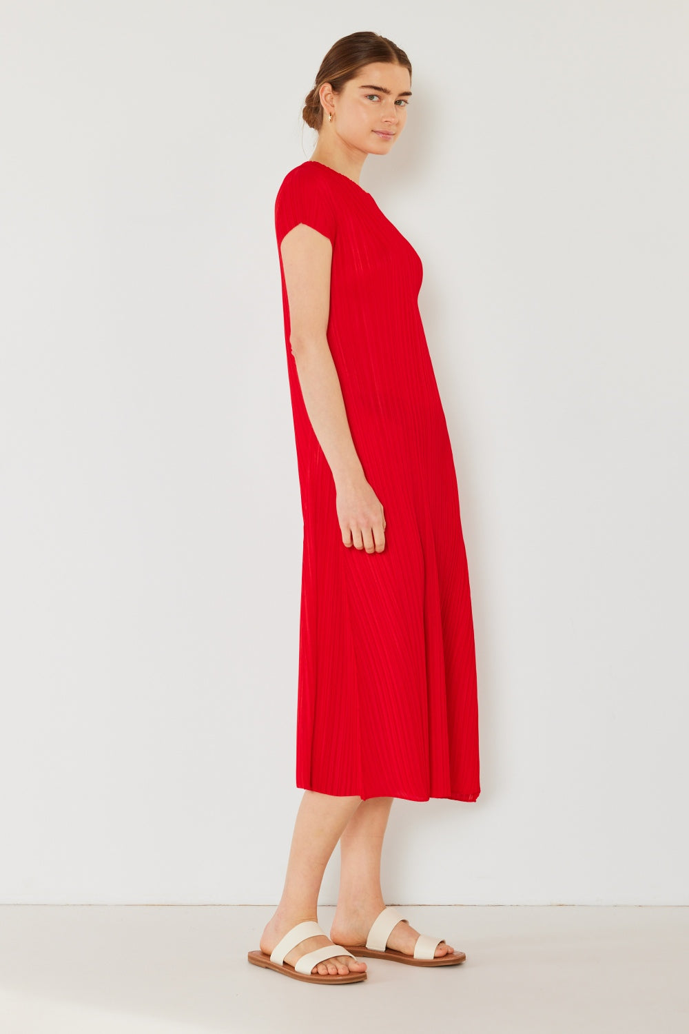 A woman exudes charm in a bright red Marina West Swim Pleated Cap Sleeve A-Line Dress, paired elegantly with white sandals and her hair neatly tied back, set against a plain white wall backdrop.