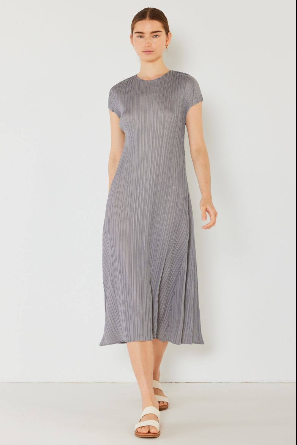 Wearing a Marina West Swim gray pleated cap sleeve A-line dress and white sandals, a person stands against a white background. Their straight hair is pulled back as they walk with a neutral expression, subtly exuding feminine charm.