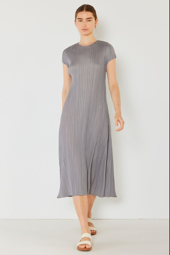 Wearing a Marina West Swim gray pleated cap sleeve A-line dress and white sandals, a person stands against a white background. Their straight hair is pulled back as they walk with a neutral expression, subtly exuding feminine charm.