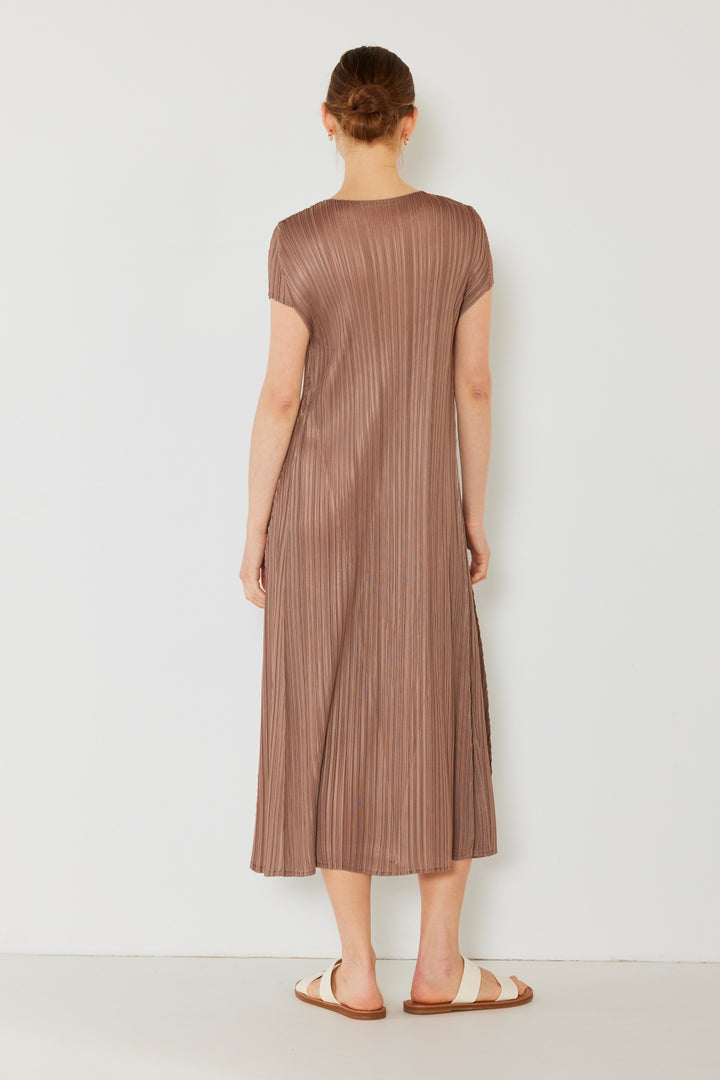 A person with tied-back hair faces a plain wall, exuding feminine charm in an ankle-length brown Marina West Swim Pleated Cap Sleeve A-Line Dress paired with sandals.