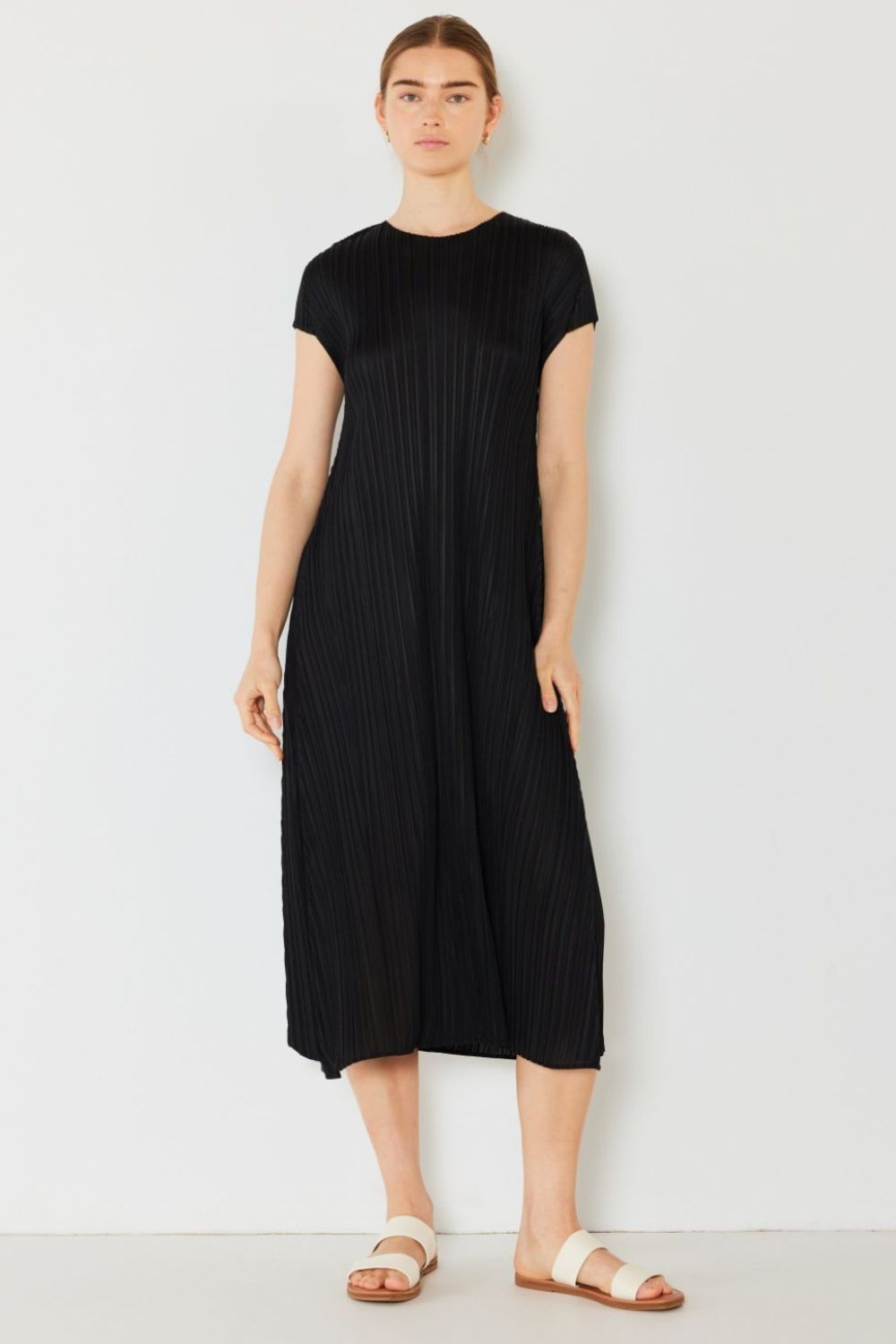 Against a white background, someone dons the Marina West Swim Pleated Cap Sleeve A-Line Dress in black paired with white sandals. With a neutral expression and arms relaxed at their sides, they exude feminine charm.