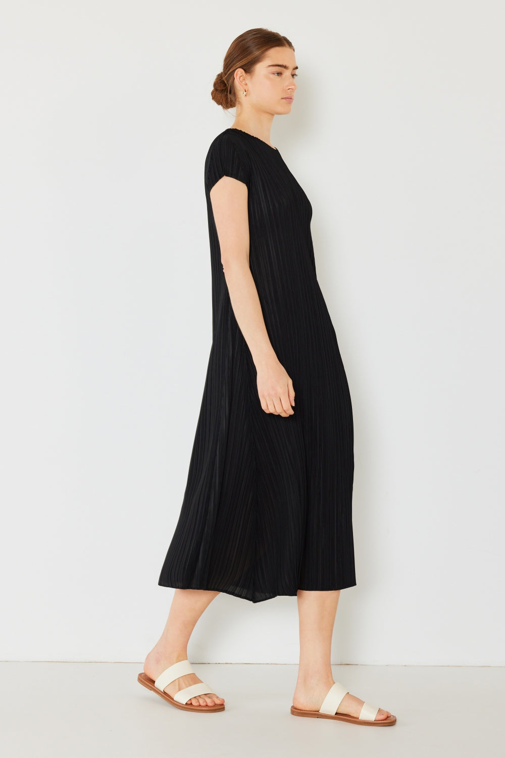 A woman with hair tied back gracefully stands sideways against a white wall, wearing the Marina West Swim Pleated Cap Sleeve A-Line Dress in black and cream sandals, exuding feminine charm.