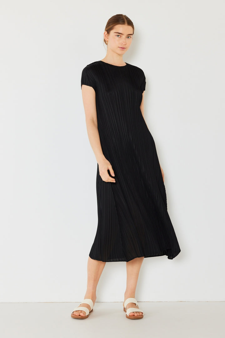 A person stands against a white background wearing a Marina West Swim Pleated Cap Sleeve A-Line Dress in black with white sandals and their hair tied back, exuding effortless feminine charm.