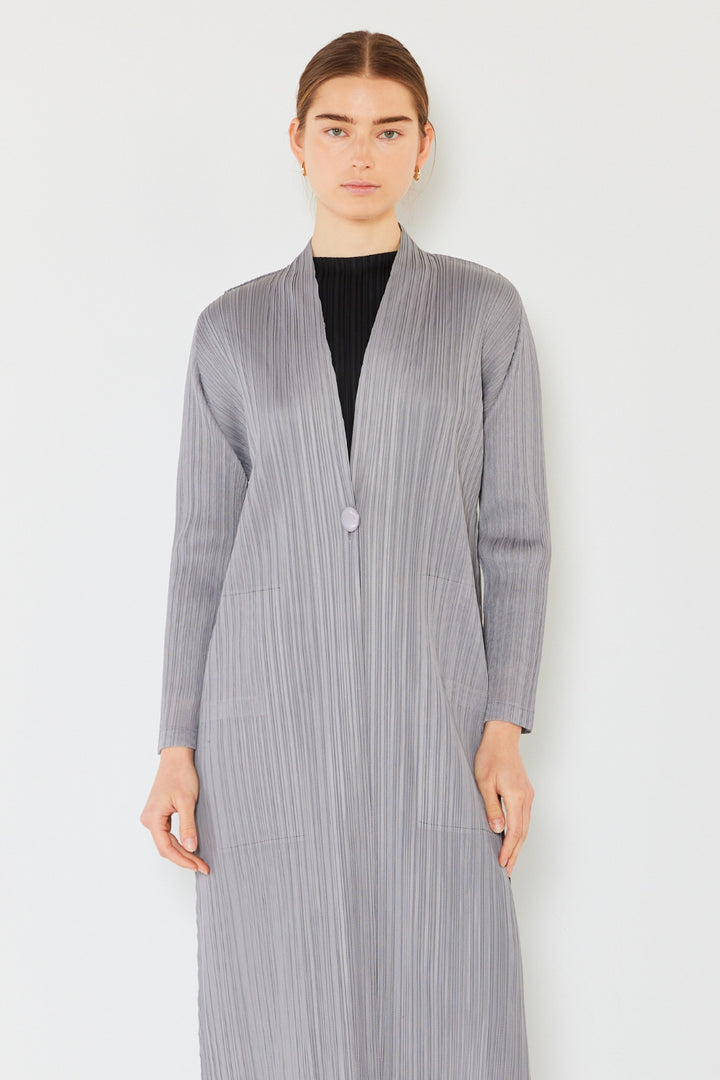 A person stands against a white background wearing the Marina West Swim Pleated Long Sleeve Cardigan in a chic, long, textured light gray design over a black top. The cardigan features a single button closure. The individual has a neutral expression and straight, slicked-back hair.