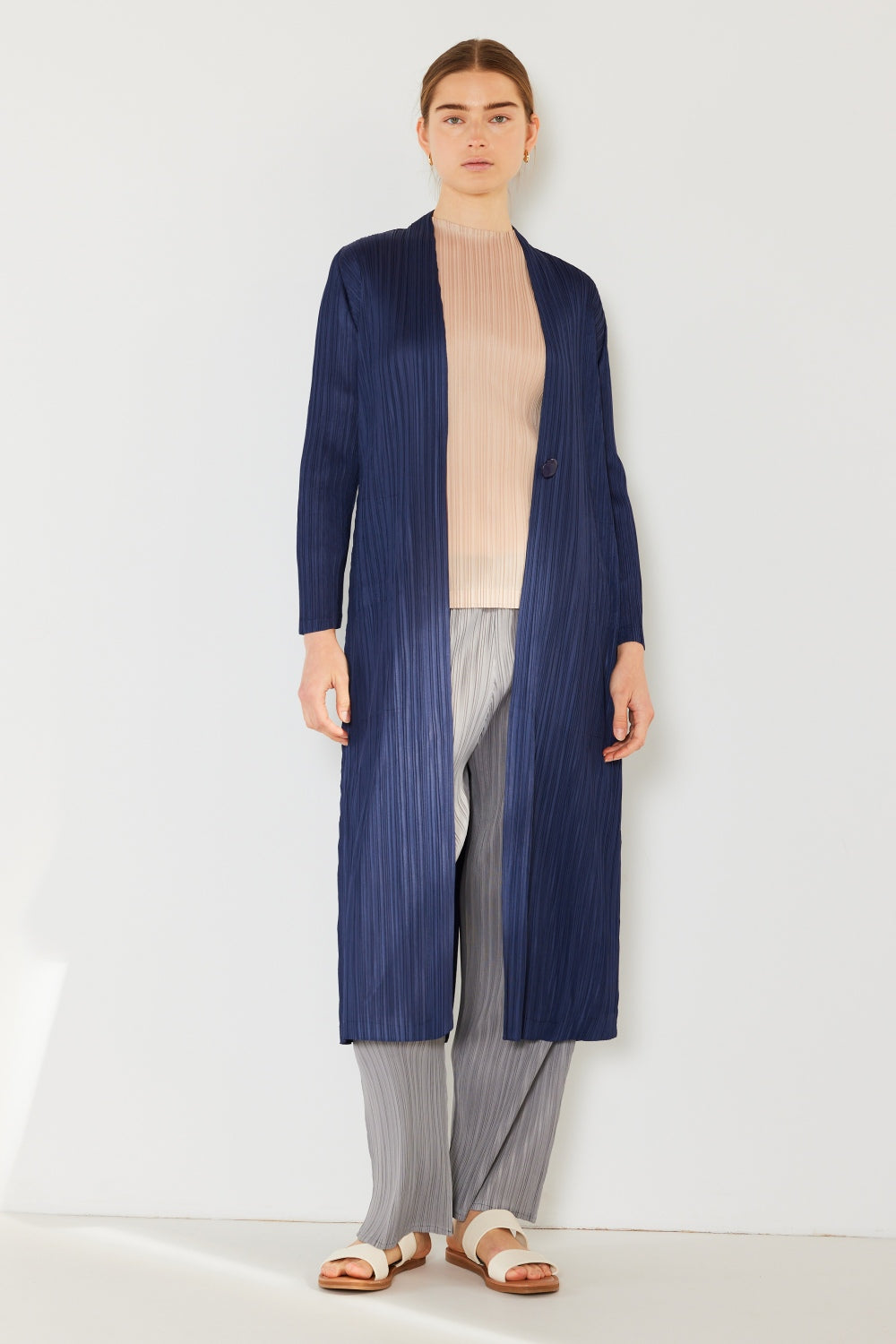 Dressed in a stylish Marina West Swim Pleated Long Sleeve Cardigan in navy blue, an individual pairs it with a light beige turtleneck, gray pleated pants, and white sandals. This versatile layering piece is showcased against a plain, light-colored background.