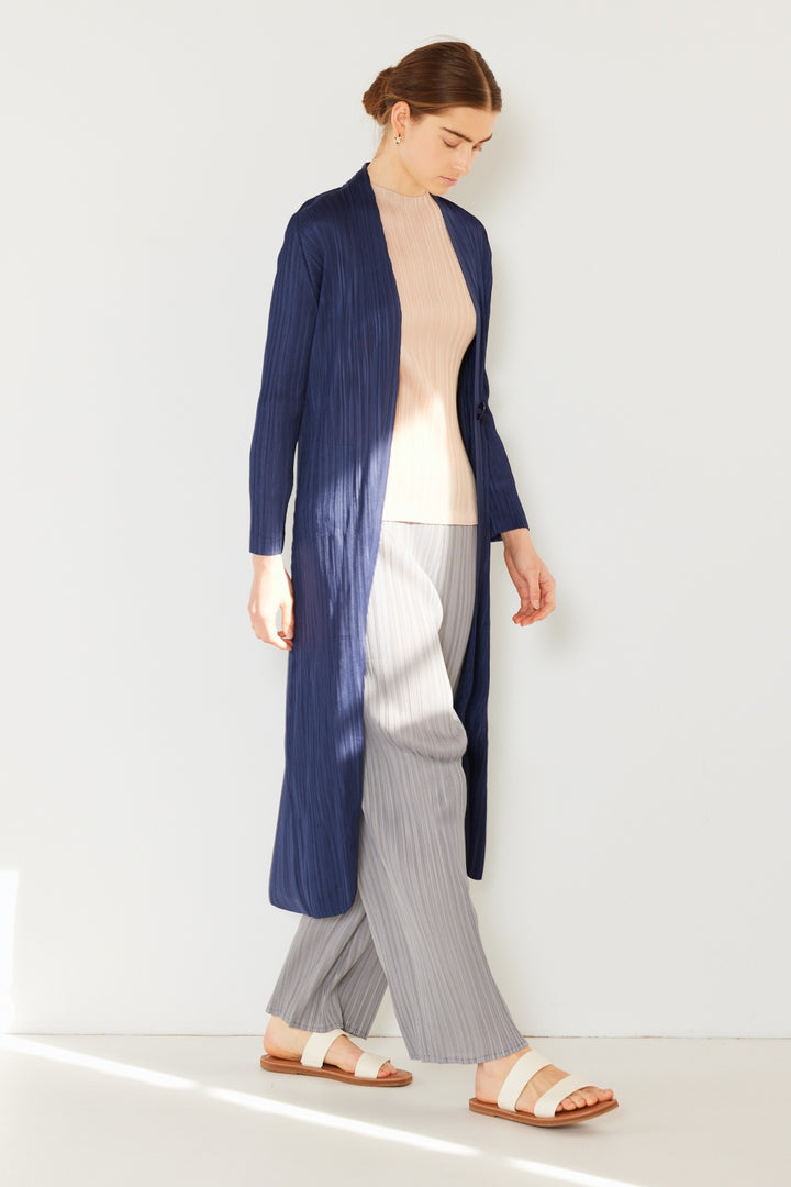 A person is standing against a white wall, wearing the Marina West Swim Pleated Long Sleeve Cardigan—a chic, long, dark blue ribbed piece that's perfect for versatile layering. Paired with a light beige top, gray pleated trousers, and cream sandals, their hair is tied back as they look down, with soft shadows dancing around them.