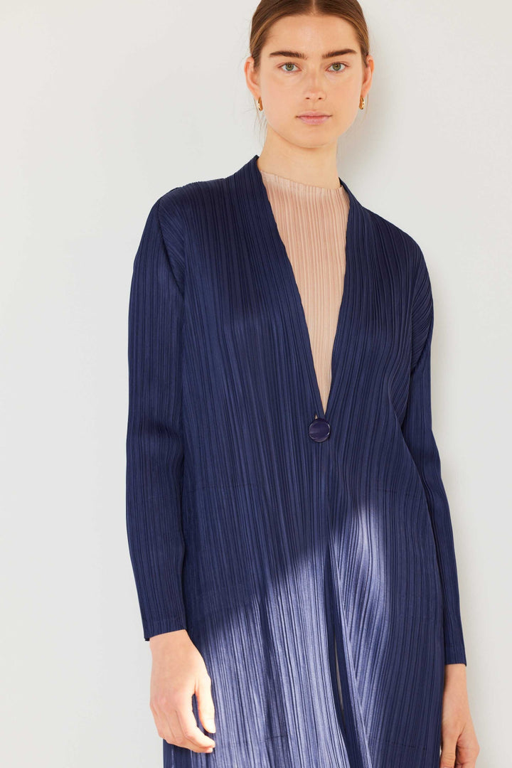 A person stands against a plain light background, gazing forward, wearing a Marina West Swim Pleated Long Sleeve Cardigan in navy blue with a single button closure over a sheer beige top. This chic ensemble serves as an elegant yet versatile layering piece suitable for any occasion.