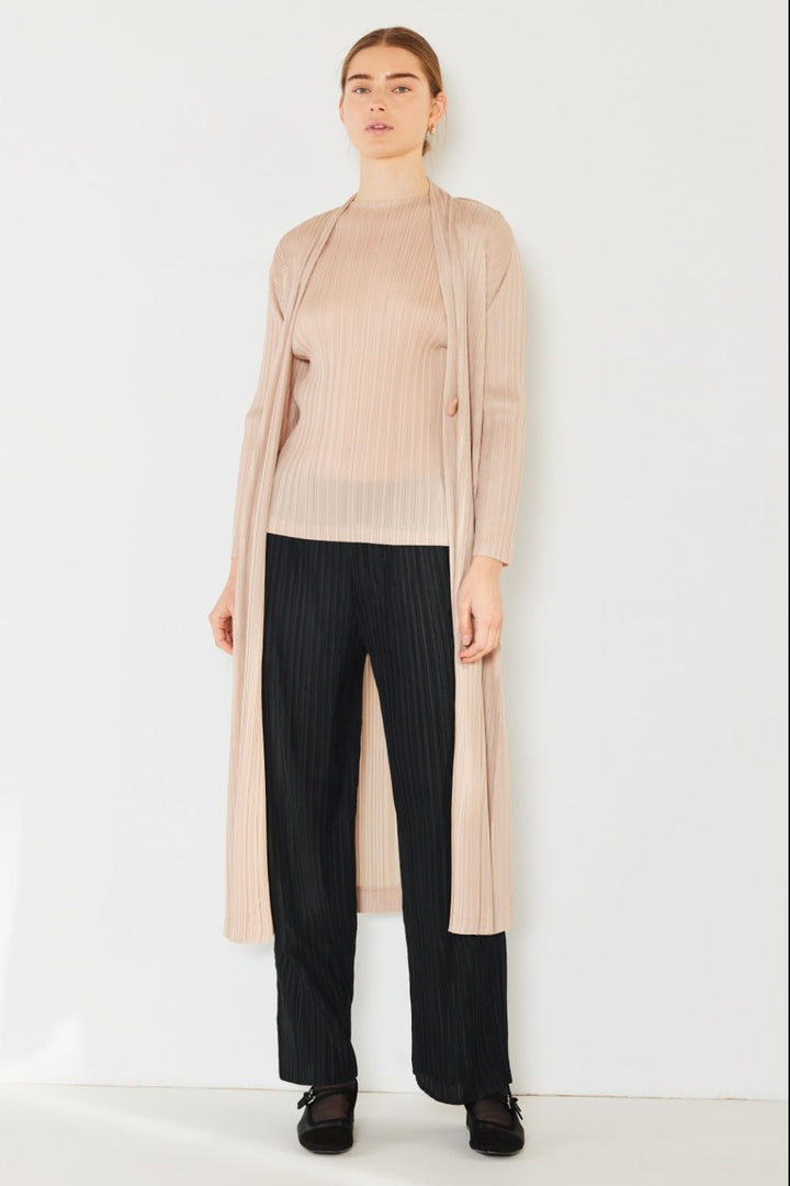 A person stands against a plain background, wearing the stylish Marina West Swim Pleated Long Sleeve Cardigan, which serves as a versatile layering piece. The light beige cardigan is draped over a matching top and black striped pants. They look directly at the camera with a neutral expression.