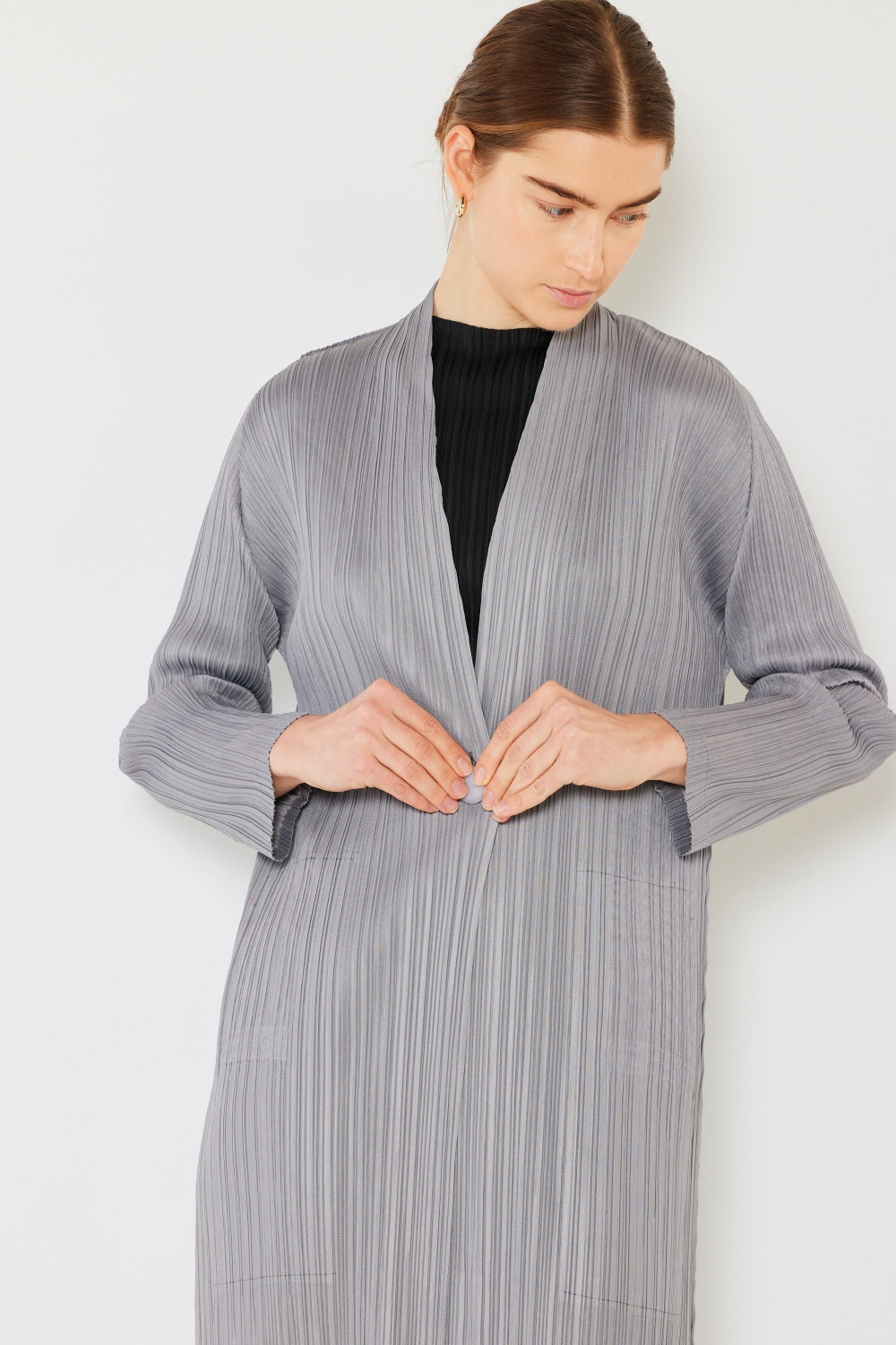 A person with brown hair pulled back is wearing the chic Marina West Swim Pleated Long Sleeve Cardigan—a long, textured, light gray coat over a black top. They are looking down slightly and touching the front of the cardigan. This versatile layering piece stands out against the plain white background.