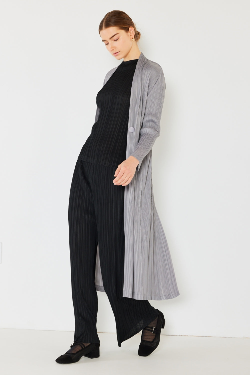 Standing against a plain white background, a person showcases their chic cardigan—a versatile layering piece. They wear the Marina West Swim Pleated Long Sleeve Cardigan in gray over a black ribbed top and matching pants, looking down while one hand gently touches the cardigan. The ensemble is completed with stylish black shoes.