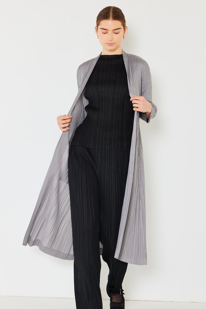 A person stands against a plain background wearing the Marina West Swim Pleated Long Sleeve Cardigan in a light grey hue, layered over a black pleated top and matching trousers. Their head is slightly tilted down, and their light brown hair is tied back.