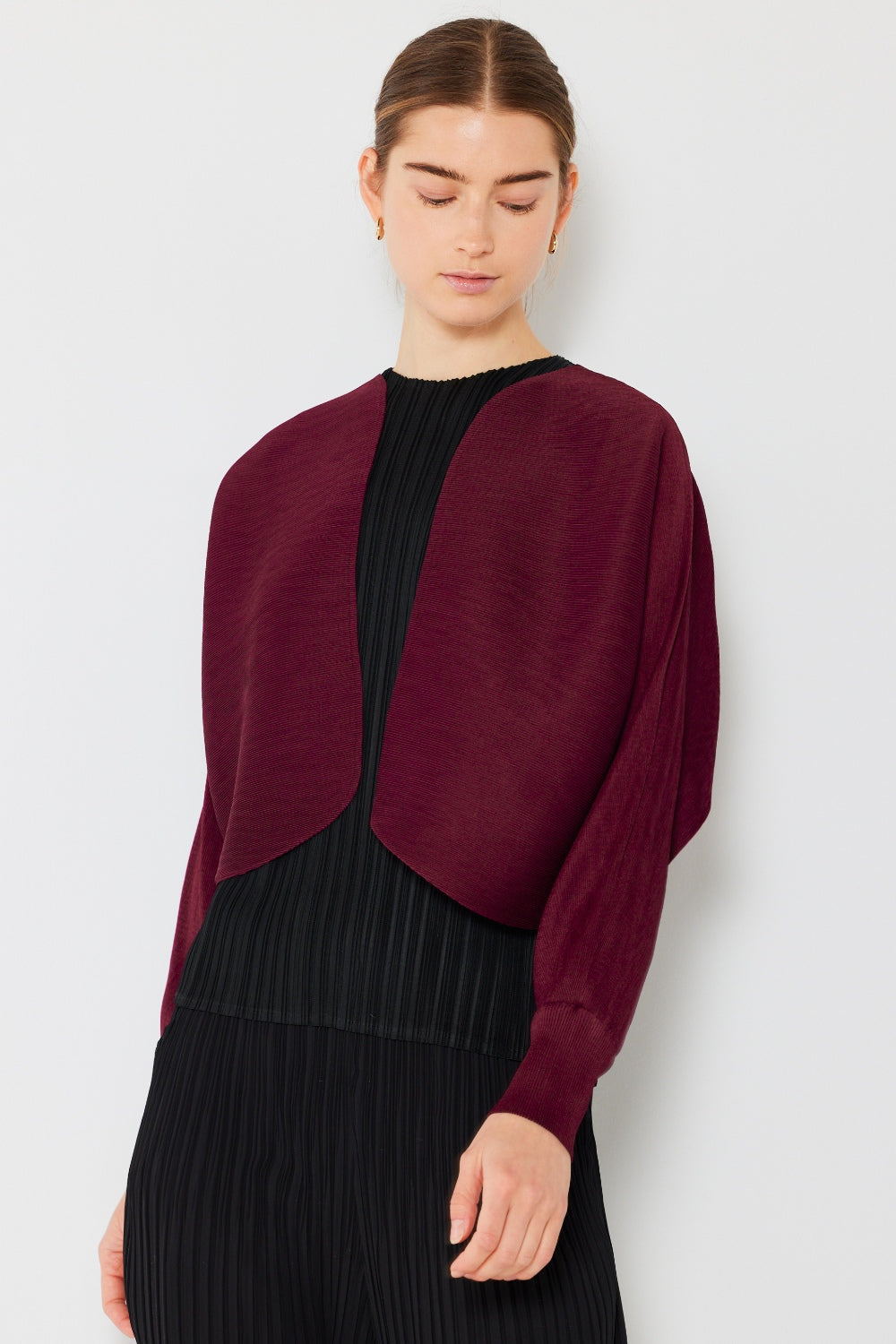 A woman with her hair tied back is wearing the Marina West Swim Rib Pleated Puff Sleeve Bolero Cardigan in burgundy over a black pleated outfit. She poses against a plain white background, exuding a feminine chic silhouette and looking downward with a calm expression.