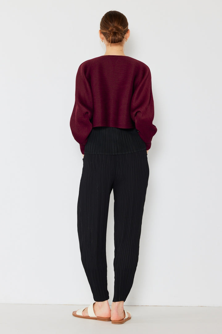 A person with brown hair tied in a bun is facing away from the camera. They are wearing a Marina West Swim Rib Pleated Puff Sleeve Bolero Cardigan over black pinstriped pants and white sandals, creating a feminine chic silhouette against the plain white wall.