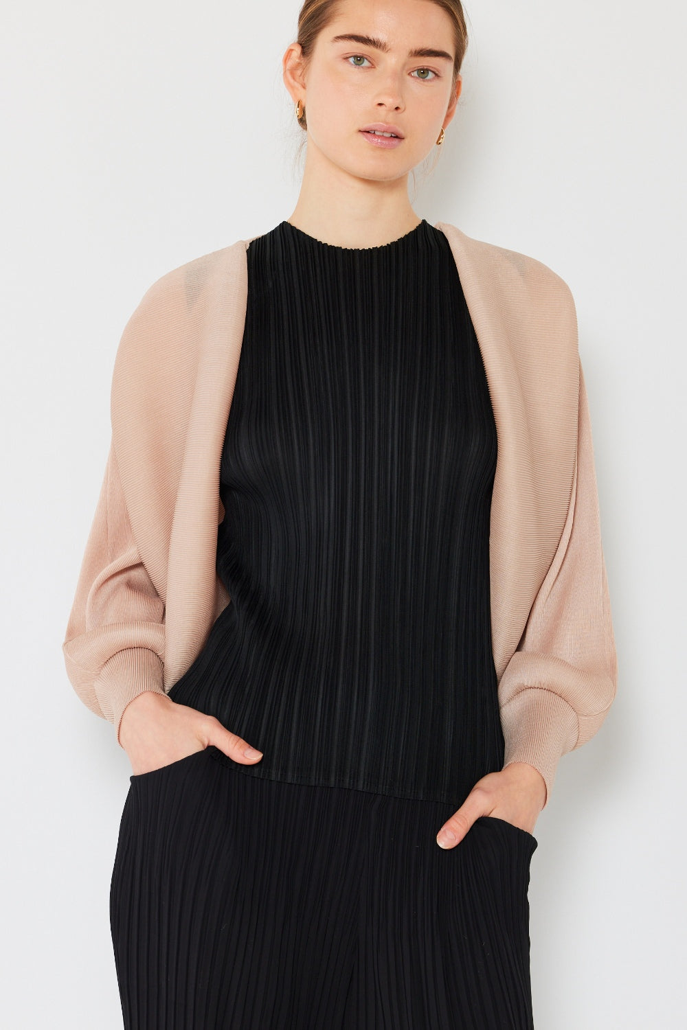 A person wearing a black pleated top and loose black pants stands against a plain light background with hands in pockets, draped in the stylish Marina West Swim Rib Pleated Puff Sleeve Bolero Cardigan over their shoulders.