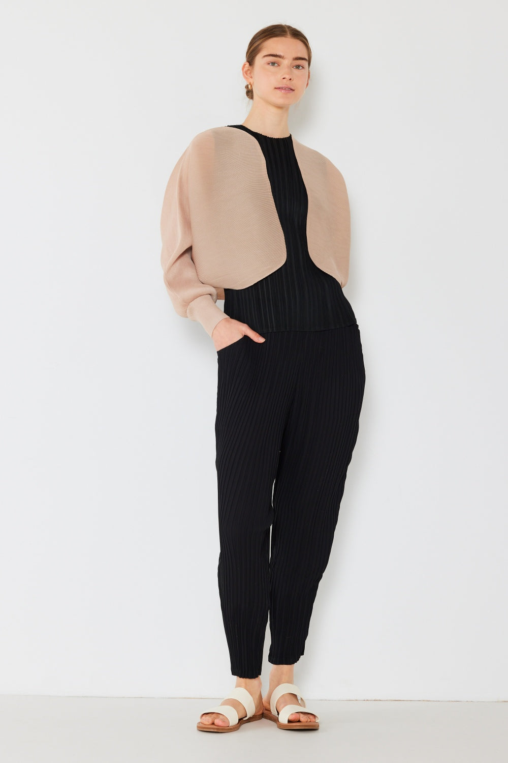 Against a white backdrop, a person models a black pleated jumpsuit, stylishly enhanced with the Marina West Swim Rib Pleated Puff Sleeve Bolero Cardigan and white sandals. Their short hair is pulled back and hands tucked into pockets, exuding an effortlessly relaxed yet feminine chic silhouette.