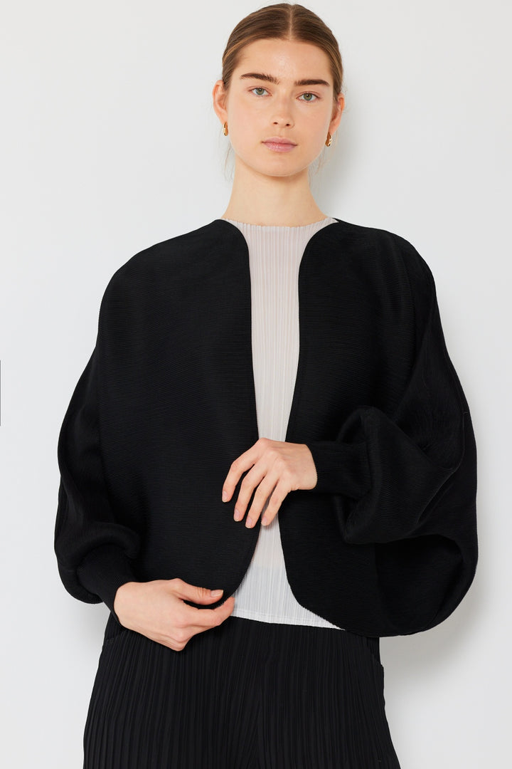 Standing against a plain background, the person with long hair wears their chic ensemble, featuring the Marina West Swim Rib Pleated Puff Sleeve Bolero Cardigan in black over a white top and pleated pants. They exude a feminine silhouette as they gaze at the camera with a neutral expression.