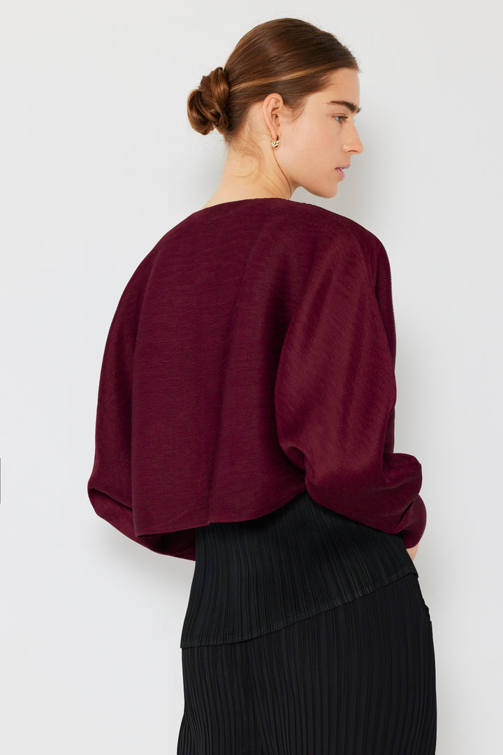 A person with brown hair in a bun is wearing the Marina West Swim Rib Pleated Puff Sleeve Bolero Cardigan in maroon, paired with a pleated black skirt, creating a chic feminine silhouette. They stand against a white background, looking to the side.