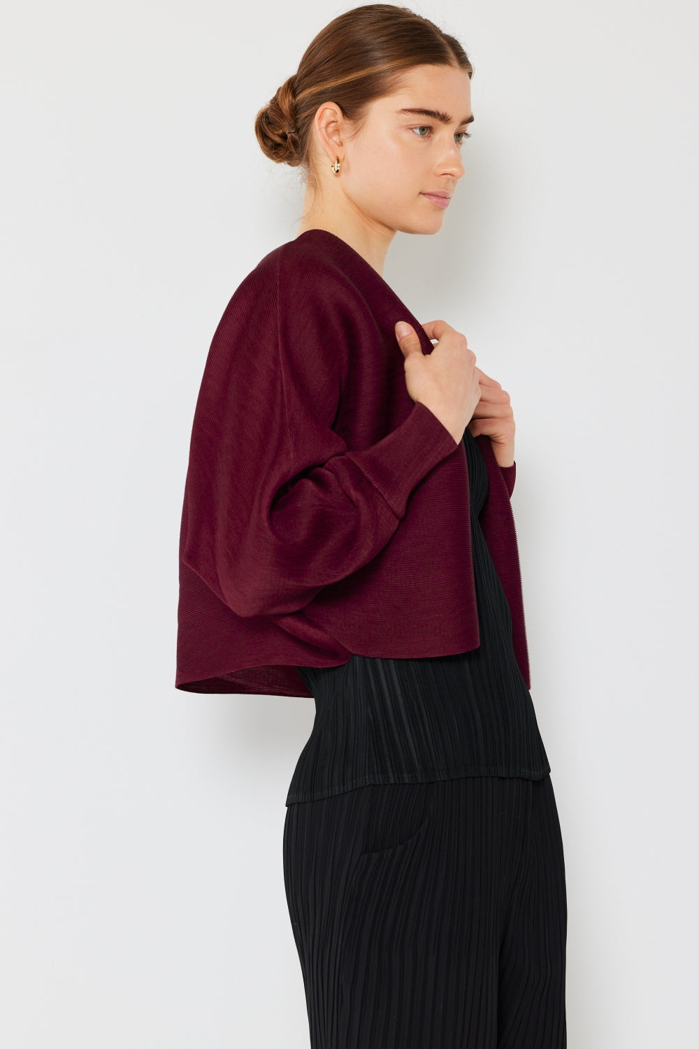 A person with brown hair is wearing the stylish Marina West Swim Rib Pleated Puff Sleeve Bolero Cardigan over a black ribbed top, exuding a feminine chic silhouette. They are standing sideways against a light gray background, perfectly highlighting the sophisticated elegance of their plum-colored ensemble.