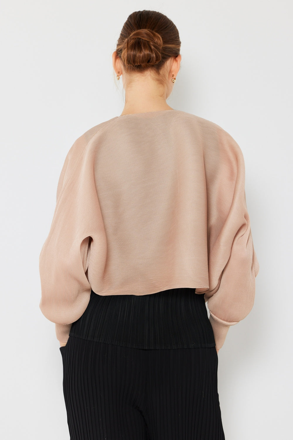 A person with their hair in a bun is standing with their back to the camera, highlighting a feminine chic silhouette while wearing the Marina West Swim Rib Pleated Puff Sleeve Bolero Cardigan in light pink, paired with black pleated pants against a plain white background.