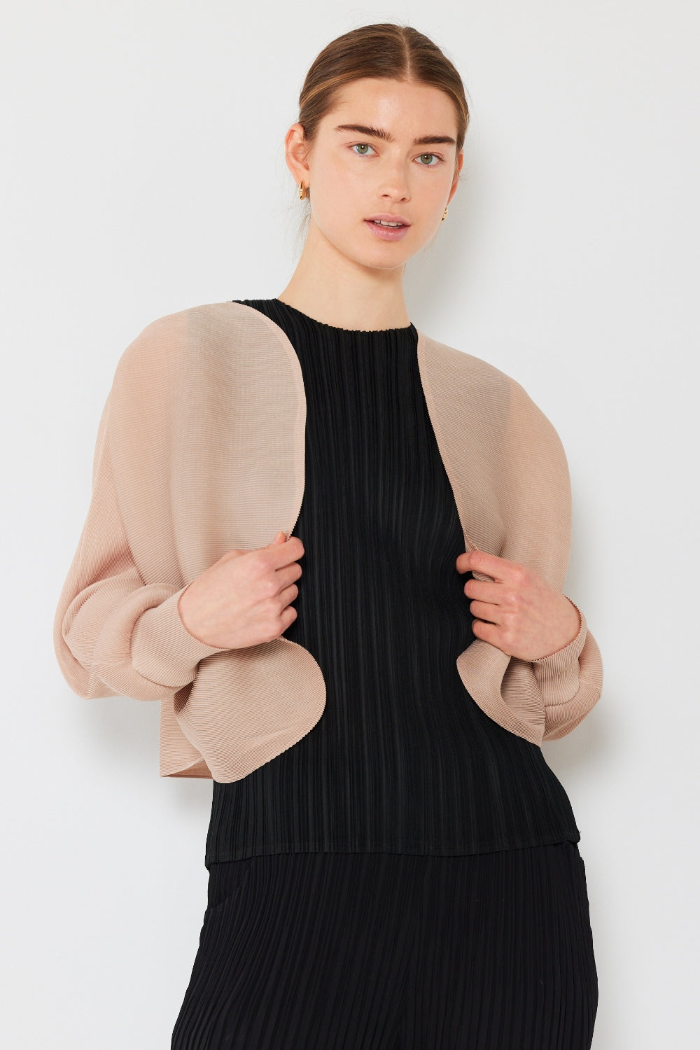 A person with brown hair is wearing a black pleated outfit paired with the Marina West Swim Rib Pleated Puff Sleeve Bolero Cardigan. The light beige, sheer garment enhances their feminine chic silhouette as they stand against a plain white background, holding the hem of the cardigan open slightly.