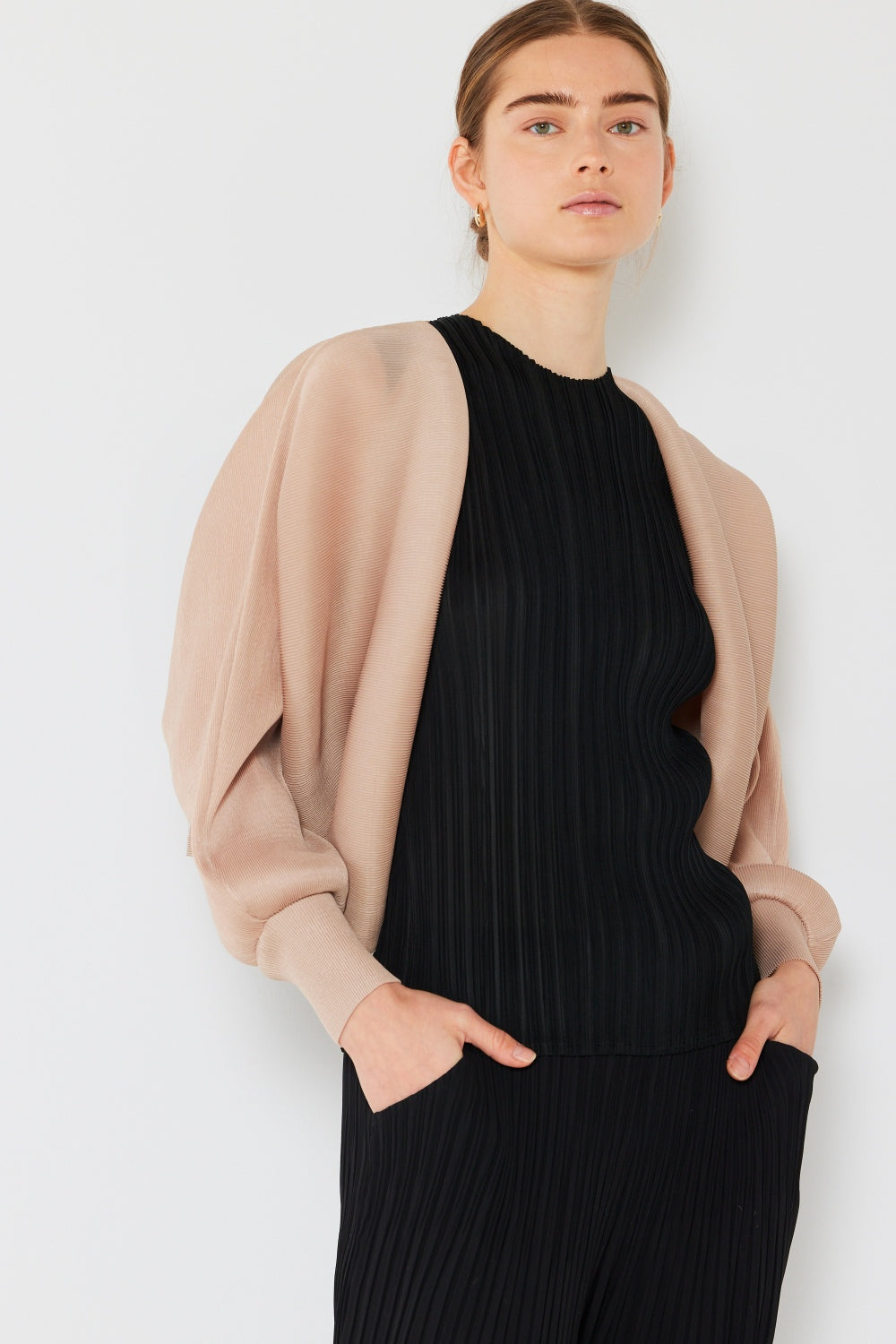 A person with brown hair is wearing a black pleated top and pants, paired with the chic Marina West Swim Rib Pleated Puff Sleeve Bolero Cardigan. They stand with hands in pockets against a plain white background, showcasing a feminine chic silhouette.