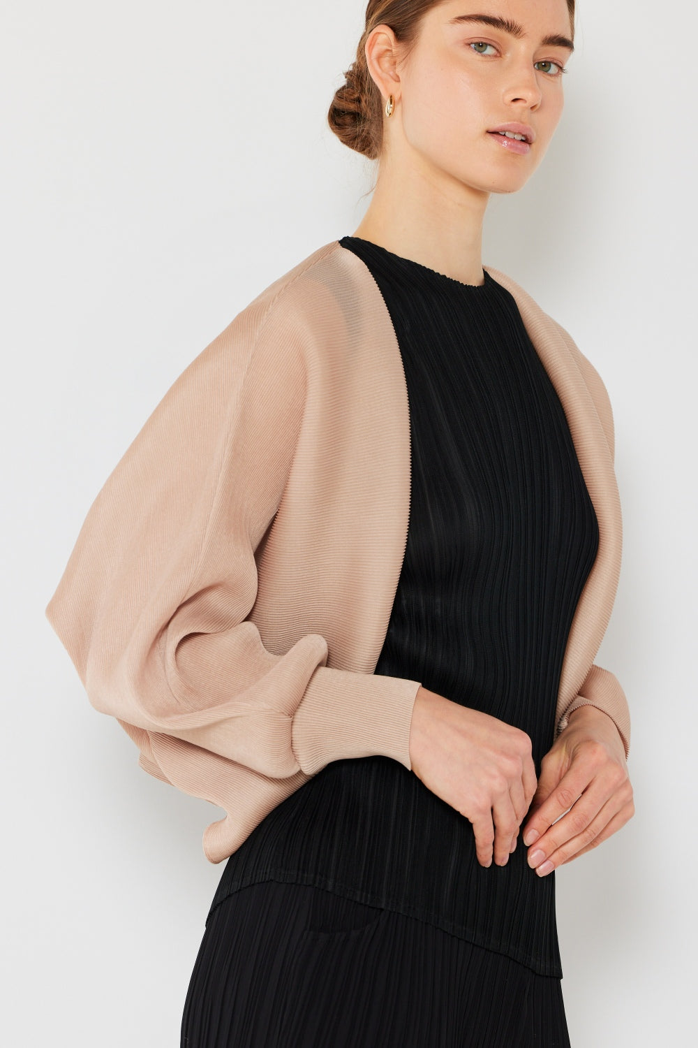 A person wearing a black pleated dress paired with the Marina West Swim Rib Pleated Puff Sleeve Bolero Cardigan stands against a plain background. The beige bolero accentuates their feminine chic silhouette. With short hair, they gaze slightly to the side.