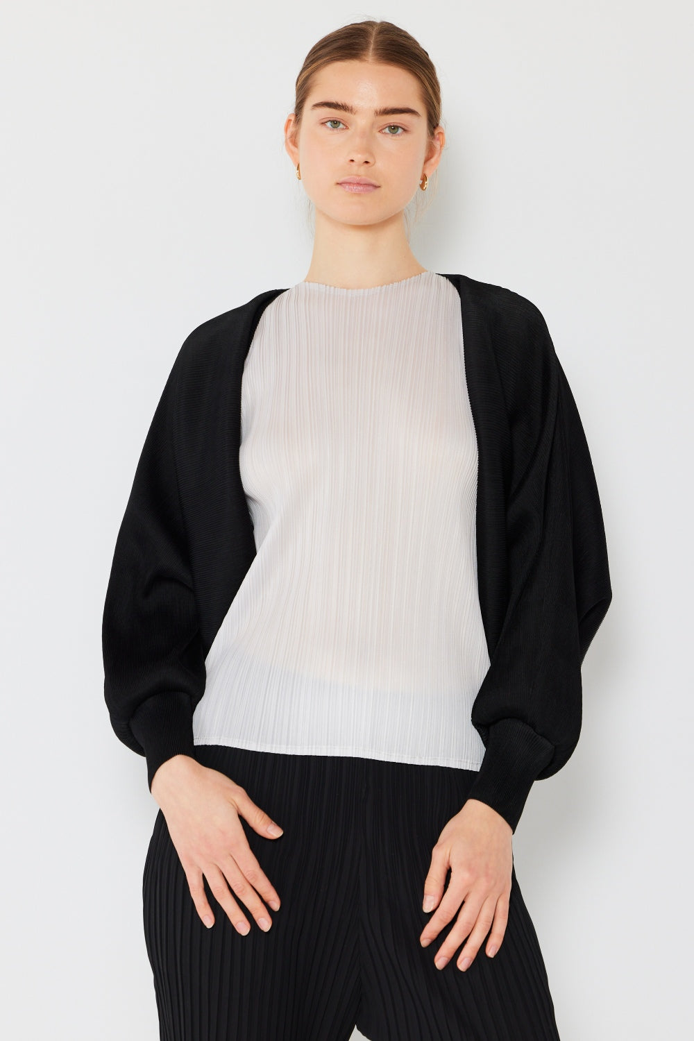 A woman stands against a light background, embodying feminine chic in a sheer pleated top paired with the stylish Marina West Swim Rib Pleated Puff Sleeve Bolero Cardigan. Her hair is tied back, with hands resting near her hips, and her expression is neutral yet elegant.