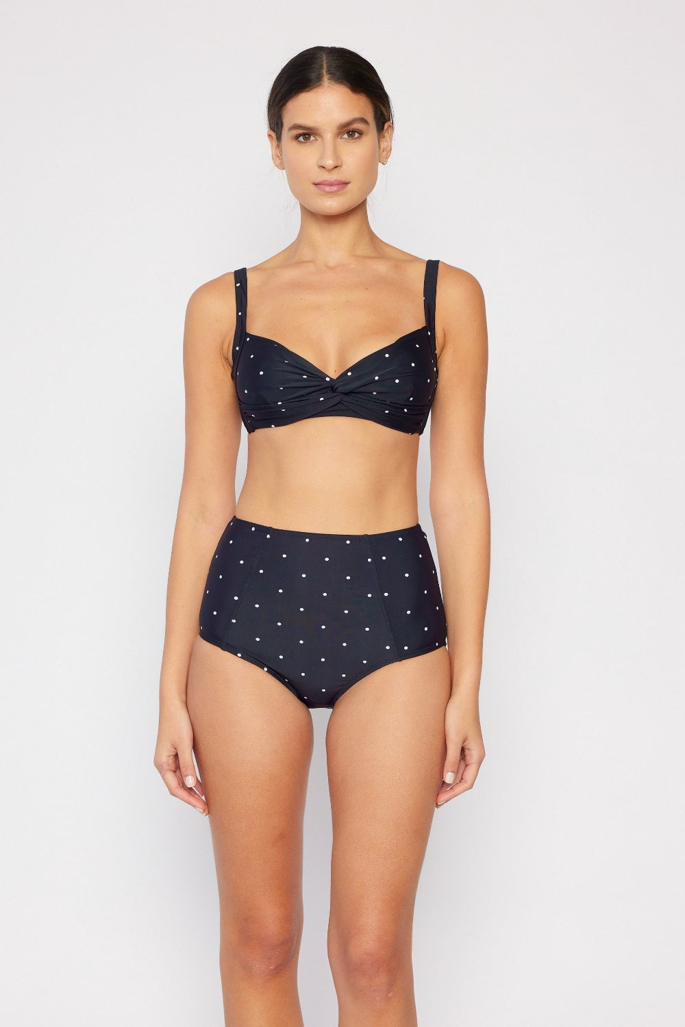 A woman stands against a plain background in a Marina West Swim Take A Dip Twisted High-Rise Bikini Set, featuring a twist-detail top and high-rise bottoms with small white polka dots. Her hair is tied back, facing forward.