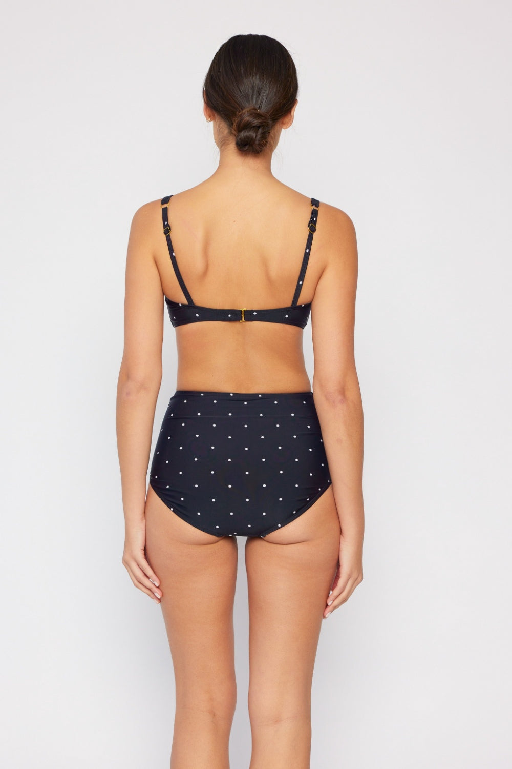 A person with dark hair in a bun models the Marina West Swim Take A Dip Twisted High-Rise Bikini Set, featuring a stylish navy blue and white polka dot design, against a plain white background, facing away from the camera.