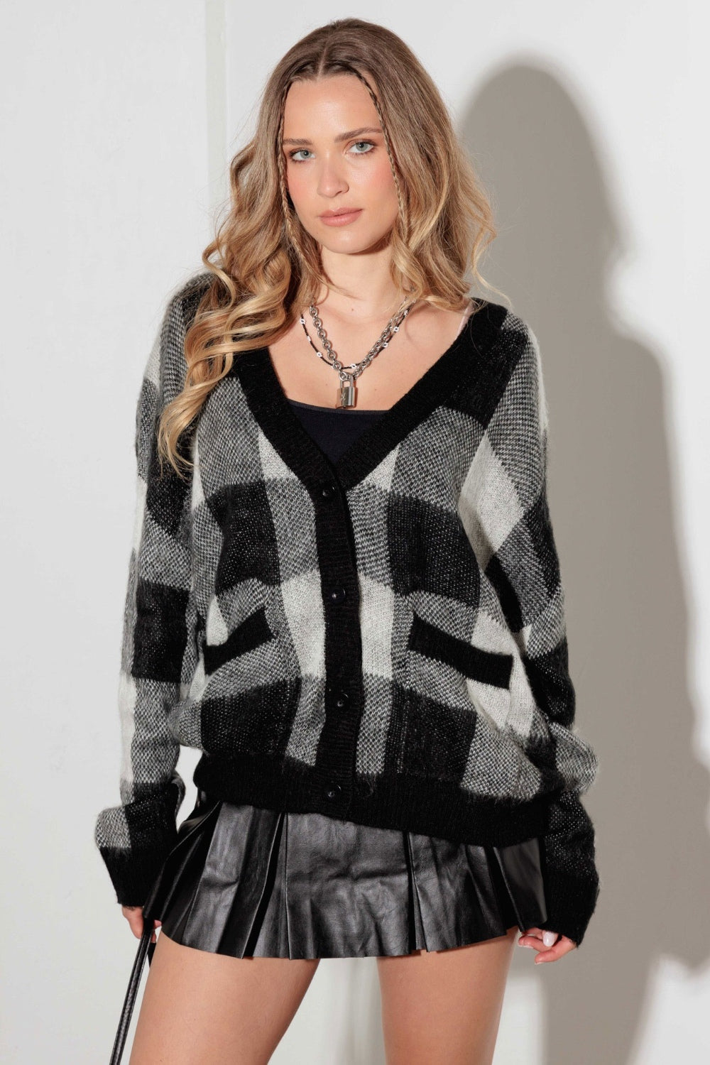 A person with long wavy hair is wearing the elegant Mustard Seed V-Neck Checkered Cardigan with Pockets in black and white over a black top, coordinated with a black pleated skirt. They stand against a white wall, hands tucked into the cardigan's pockets, highlighting this versatile wardrobe staple.