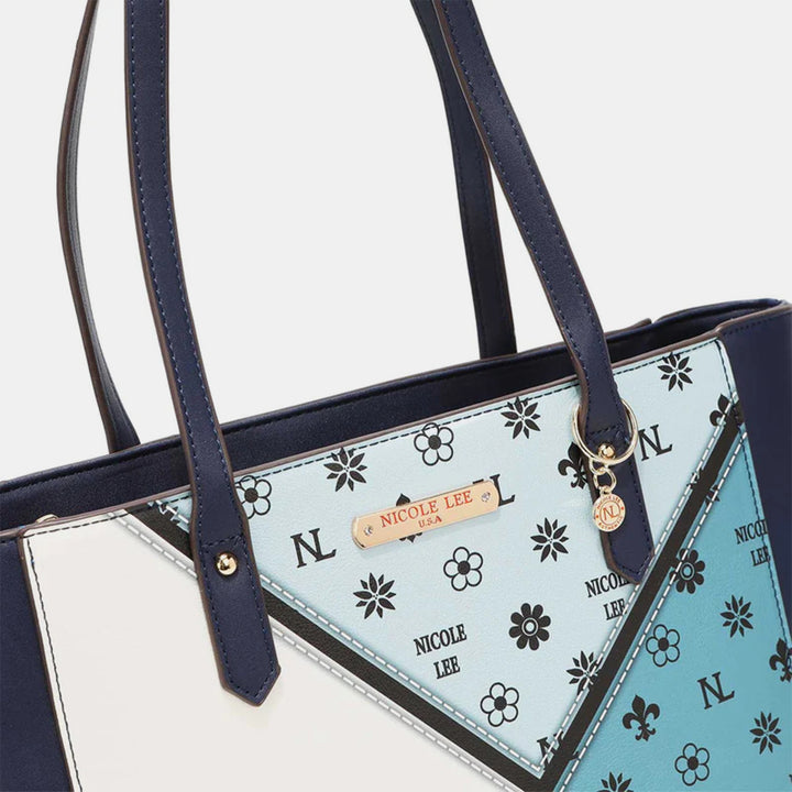 The Nicole Lee USA 3-Piece Color Block Handbag Set is a stylish vegan leather accessory with dark blue straps and a geometric design, featuring white, light blue, and darker blue sections adorned with floral patterns. It includes a gold-toned charm and an engraved metal label.