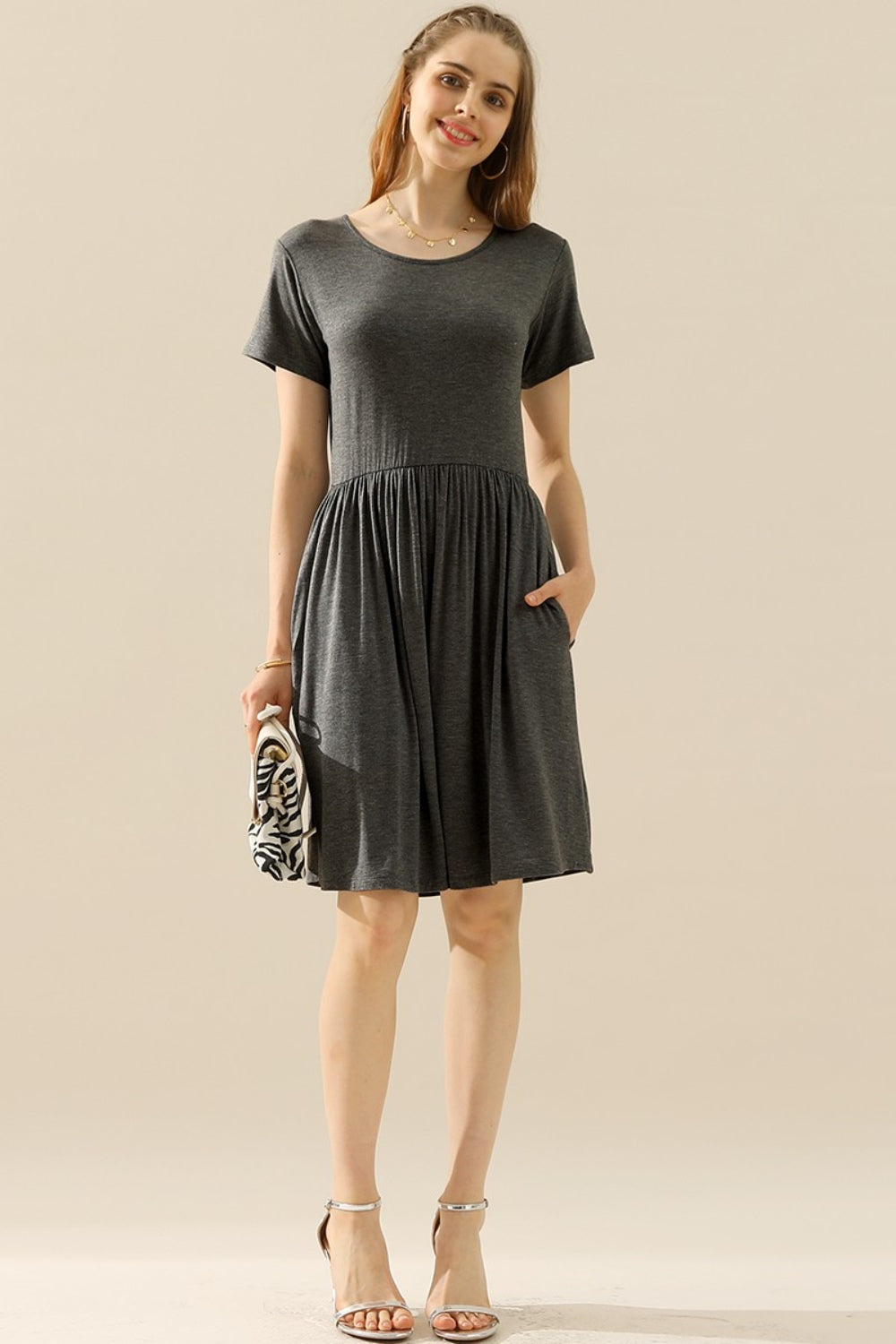 A woman stands smiling in the Ninexis Full Size Round Neck Ruched Dress with Pockets, its knee-length gray fabric elegantly gathered at the waist. She holds a patterned clutch in one hand and wears nude heels against a neutral beige background.