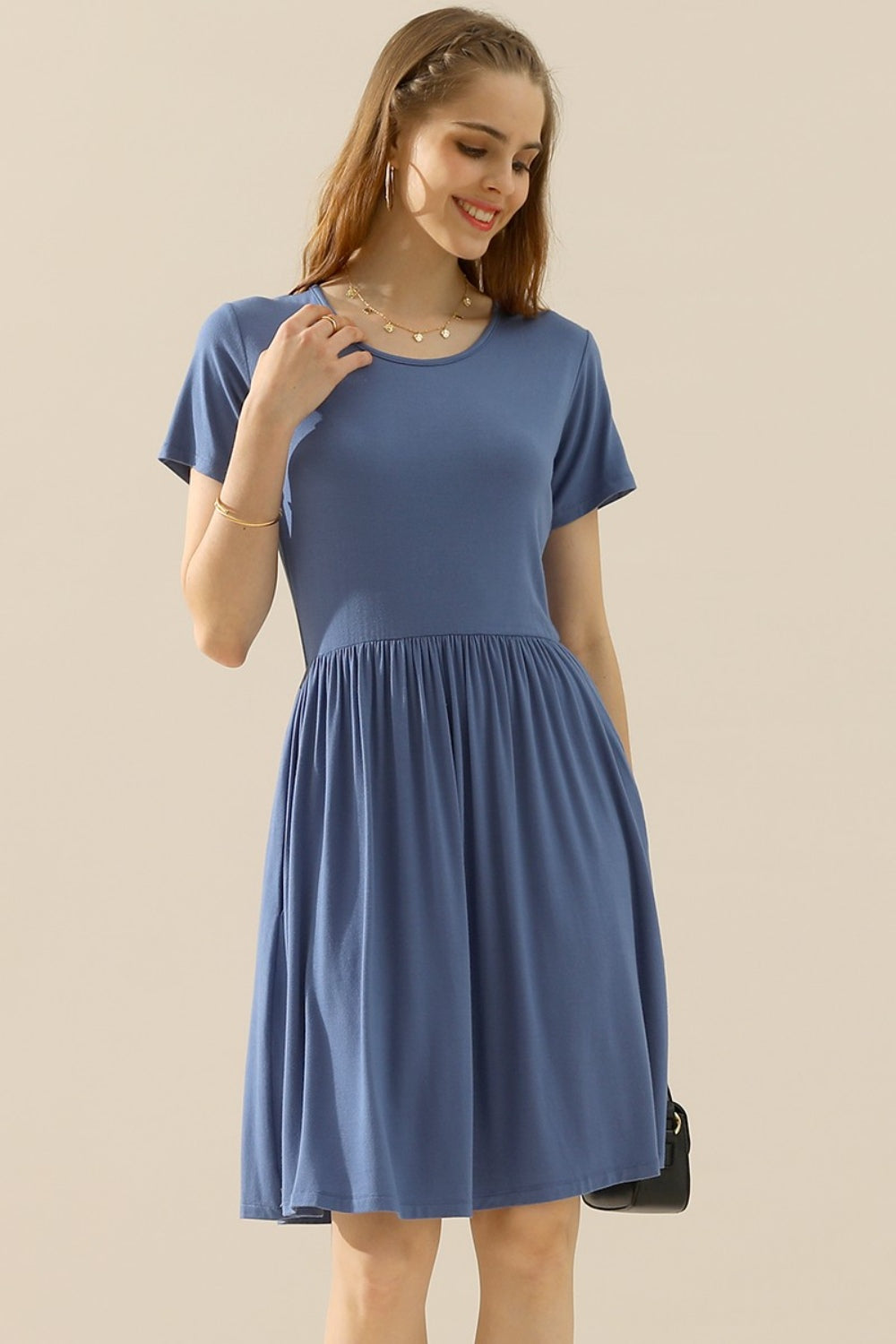 A woman wearing the Ninexis Full Size Round Neck Ruched Dress with Pockets stands smiling, her hand touching her hair. She accessorizes with a necklace, earrings, and a bracelet while holding a small black bag. The casual blue dress complements the plain beige background.