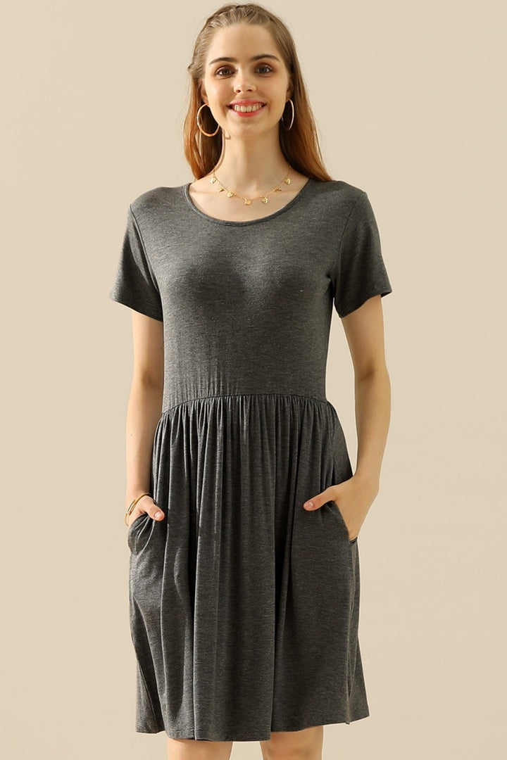 A woman beams in a versatile Ninexis Full Size Round Neck Ruched Dress with Pockets. She has long hair and stylishly pairs her look with hoop earrings and a necklace, against a simple backdrop that keeps all attention on her chic ensemble.