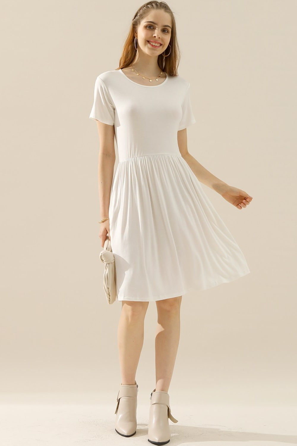 A woman is posing in a Ninexis Full Size Round Neck Ruched Dress with short sleeves and discreet pockets, holding a small clutch. The dress perfectly complements her light-colored ankle boots and long hair as she stands against a neutral beige background.