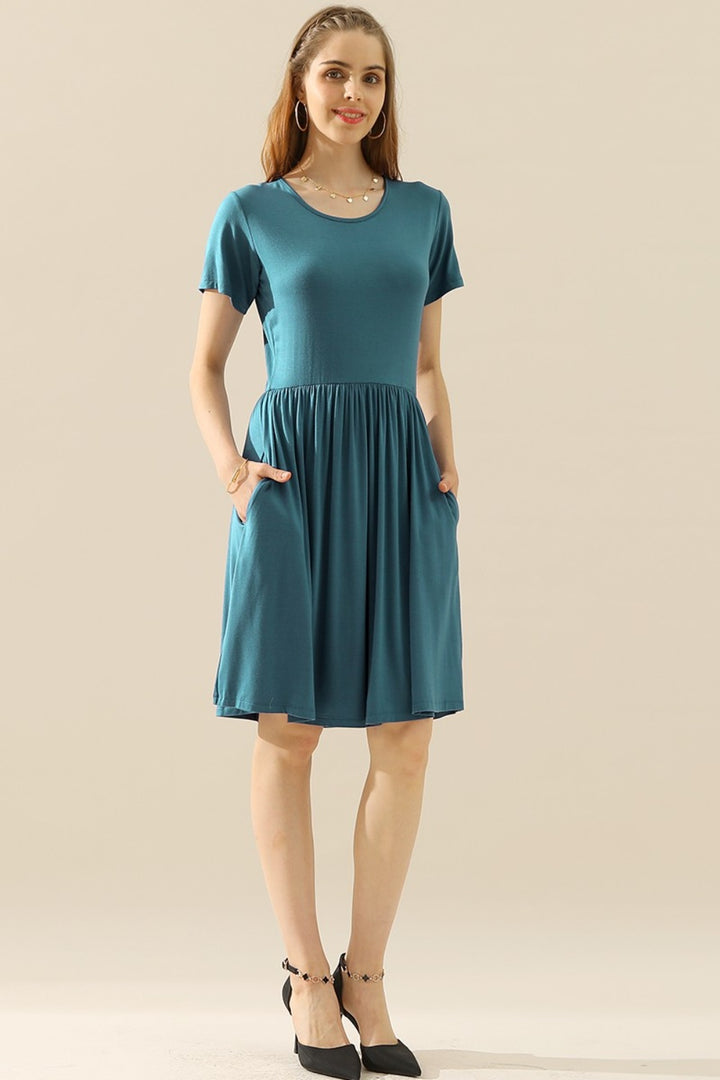 A woman poses against a plain background in a versatile, knee-length teal dress with short sleeves and pockets, identified as the Ninexis Full Size Round Neck Ruched Dress with Pockets. The ensemble is completed with black ankle-strap shoes and hoop earrings. Her hands rest in the pockets as her hair flows freely down.