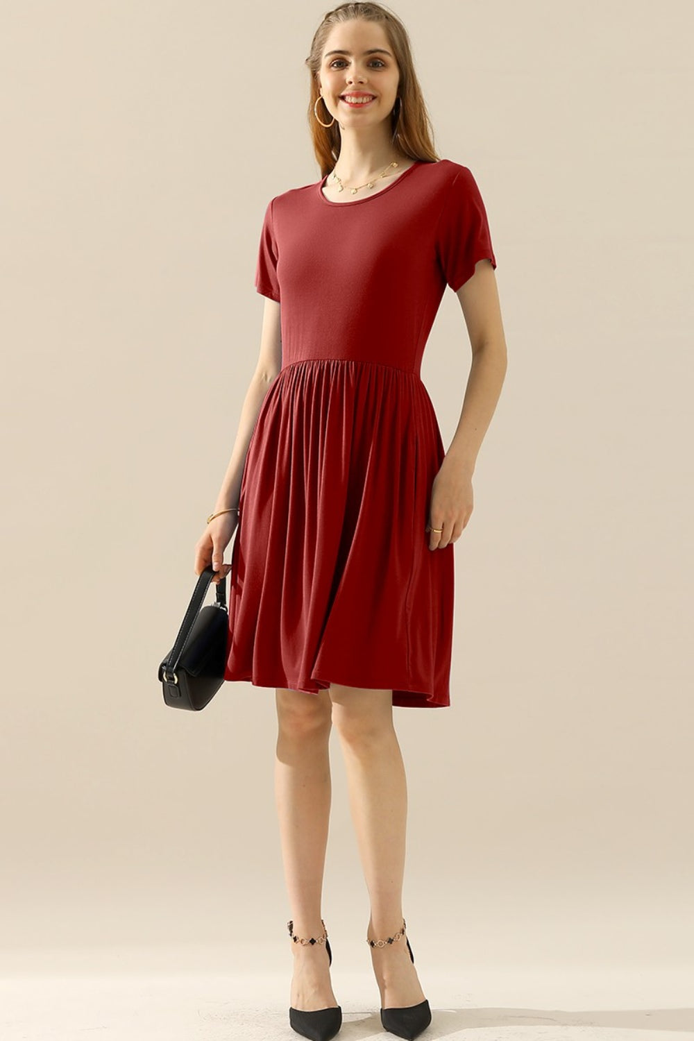 A woman wears the Ninexis Full Size Round Neck Ruched Dress with Pockets, featuring a short sleeve and knee-length finish in red. She holds a black handbag while smiling softly. Her comfortable, stretchy fabric dress is complemented by handy pockets, and she pairs it with black footwear adorned with ankle straps against a plain light beige background.