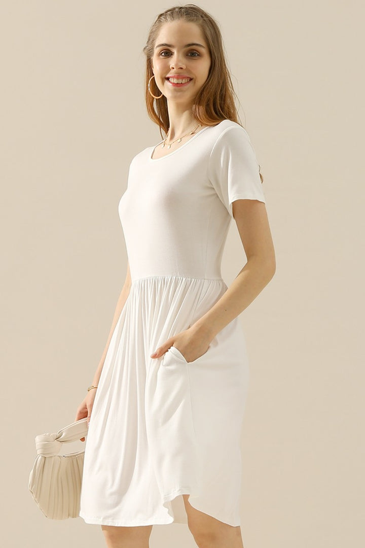 A woman in a Ninexis Full Size Round Neck Ruched Dress with Pockets is smiling while standing against a plain background. Her long hair cascades over the shoulders of her chic outfit, and she is holding a white purse.