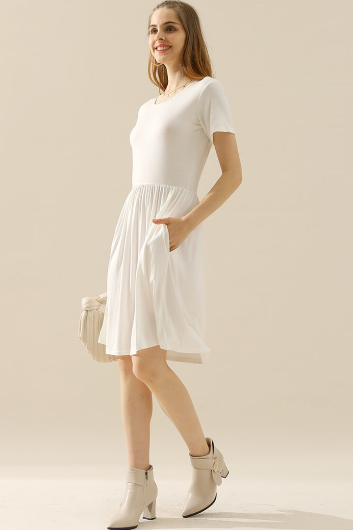 A woman stands wearing the Ninexis Full Size Round Neck Ruched Dress with Pockets, a versatile piece featuring a flowing skirt. She holds a light-colored handbag and smiles against a neutral background, while her beige ankle boots complete the look.