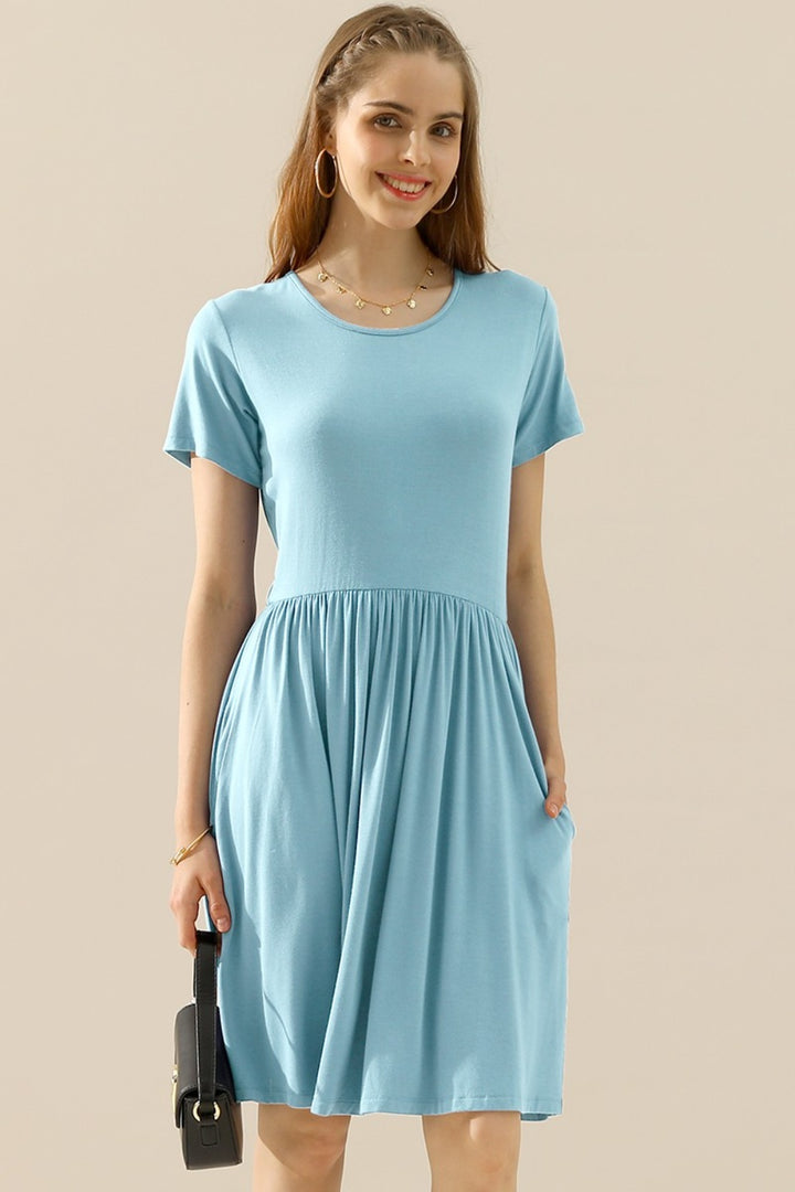 A woman wearing a versatile Ninexis Full Size Round Neck Ruched Dress in light blue, featuring a gathered waist and convenient pockets, smiles while holding a small black purse. She has long hair, hoop earrings, and wears a necklace against a neutral background.