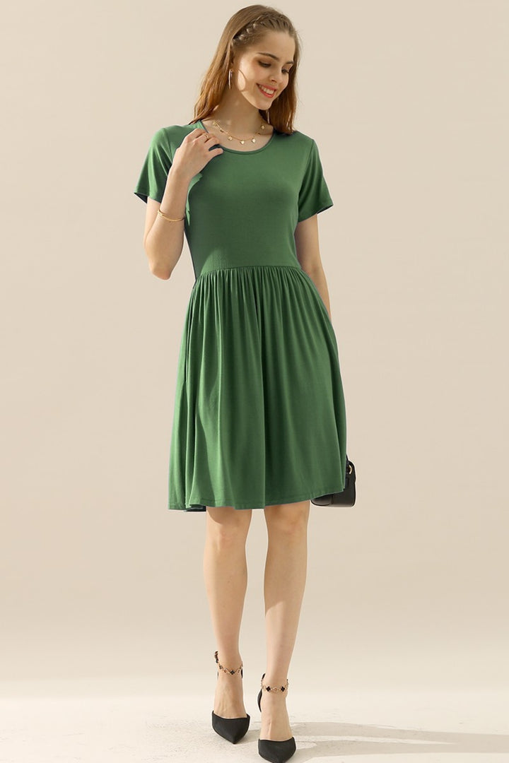 A person is smiling against a neutral background, wearing the Ninexis Full Size Round Neck Ruched Dress with Pockets, a knee-length green dress featuring short sleeves and a gathered waist. Their shoulder-length hair complements the dress's versatile design.