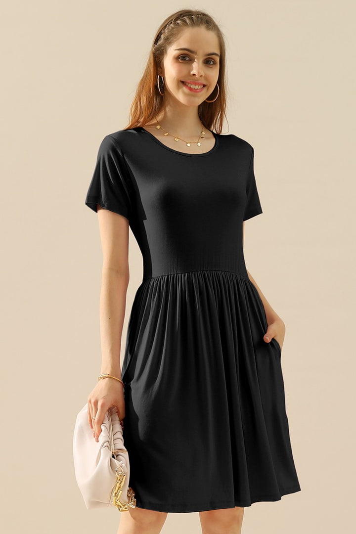 A woman in a stylish black Ninexis Full Size Round Neck Ruched Dress with Pockets stands smiling while holding a light pink handbag. She accessorizes with hoop earrings and a delicate necklace, and her hair is partially braided. The dress features short sleeves and a gathered waist.