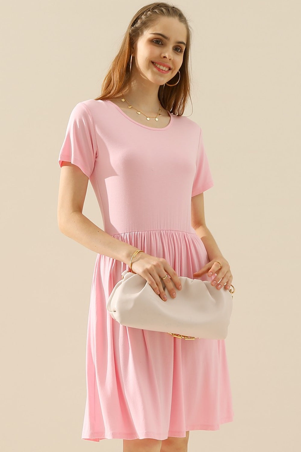 A woman stands against a plain background wearing a Ninexis Full Size Round Neck Ruched Dress with Pockets in light pink. She holds a beige clutch bag and smiles, her braided hair cascading on one side. Accessorizing with a necklace and bracelet, she showcases the versatile elegance of her outfit.