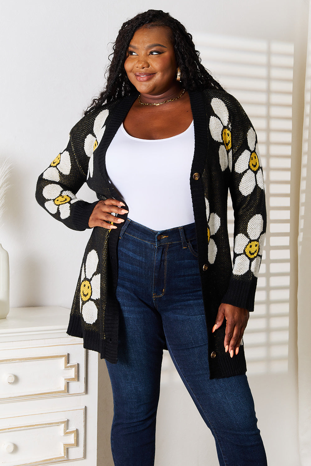 The woman is wearing the Perfee Floral Button Down Longline Cardigan, featuring a charming floral pattern of white daisies and smiley faces. She pairs it with a white top and blue jeans and stands beside a white dresser in a sunlit room, smiling and gazing to the side.