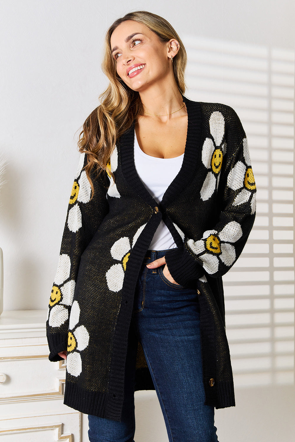 A woman with long hair smiles while wearing the Perfee Floral Button Down Longline Cardigan, showcasing a playful floral pattern of large white daisies, each with a yellow smiley face at the center. She stands indoors near a white piece of furniture, with light streaming in through a window.