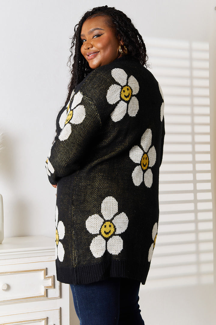 A woman with long curly hair is standing sideways, smiling at the camera. She is wearing the Perfee Floral Button Down Longline Cardigan over dark jeans in a bright room with a white dresser.